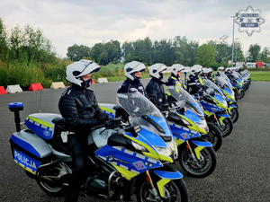 na zdjęciu widoczni usytuowani na placu manewrowym, w szeregu policjanci w czarnych kombinezonach motocyklowych i białych kaskach, siedzący na oznakowanych motocyklach służbowych z napisem policja