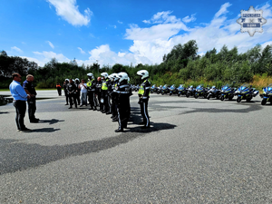na zdjęciu widoczni stojący na placu manewrowym, w szeregu policjanci w czarnych kombinezonach motocyklowych i białych kaskach, przed nimi dwaj instruktorzy policyjni , na drugim planie zaparkowane w rzędzie oznakowane motocykle służbowe z napisem policja