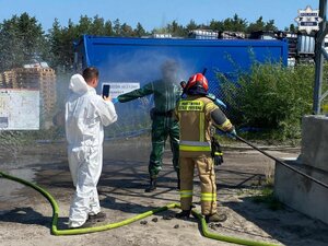 Strażak spłukuje kombinezon ochronny na osobie, która ma maskę gazową. Obok stoi mężczyzna w kombinezonie ochronnym, który rejestruje sytuacje telefonem komórkowym.