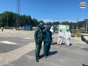 Dwie osoby w zielonych kombinezonach ochronnych i maskach gazowych. Dalej stoją trzy osoby w białych kombinezonach ochronnych i maskach gazowych.