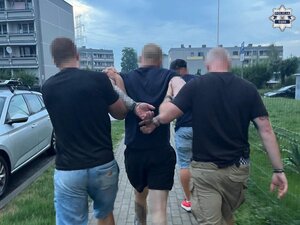 Nieumundurowani policjanci prowadzą zatrzymanego mężczyznę, który ma założone kajdanki na ręce trzymane z tyłu.
