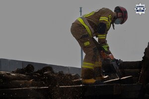 Strażak przecina piłą drewnianą belkę, składowaną w kontenerze.