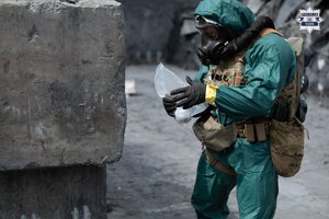 Osoba w kombinezonie ochronnym i masce gazowej zabezpiecza substancję.