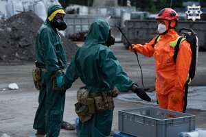 Dwie osoby w kombinezonach ochronnych i maskach gazowych są spłukiwane przez trzecią osobę, która także jest w kombinezonie ochronnym, kasku ochronnym i maseczce na twarzy.