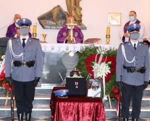 Zdjęcie przedstawia dwóch policjantów z warty honorowej w kościele przy urnie, upamiętniającej fotografii oraz odznaczenia  zmarłego policjanta. W oddali znajdują się księża prowadzący ceremonię.