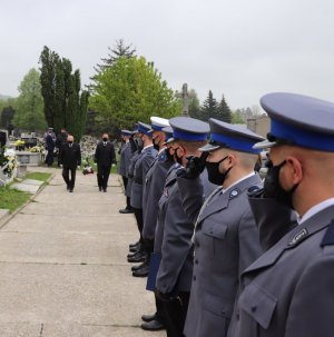 Zdjęcie przedstawia pracowników zakładu pogrzebowego niosących urnę oraz salutujących policjantów.