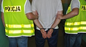 Zdjęcie przedstawia dwóch policjantów w kamizelkach odblaskowych z napisem &quot;POLICJA&quot;, którzy trzymają zatrzymanego mężczyznę. Mężczyzna ma założone kajdanki na ręce trzymane z tyłu.