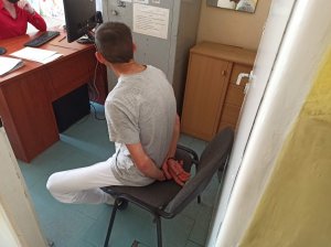 Zatrzymany mężczyzna siedzi na krześle w pokoju podczas prowadzenia z nim czynności przez pracownika pionu kryminalnego. Mężczyzna siedzi i ma założone kajdanki na ręce trzymane z tyłu.