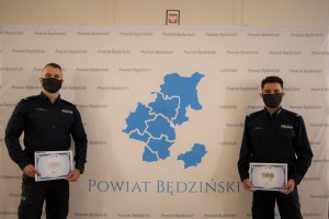 Dwaj umundurowani policjanci stojący obok siebie. W tle widoczna tablica z napisem Powiat Będziński oraz Godło Polski.
