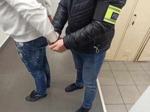 Policjant po cywilnemu z opaską z napisem Policja, zakładający kajdanki na ręce trzymane z tyłu zatrzymanemu mężczyźnie.