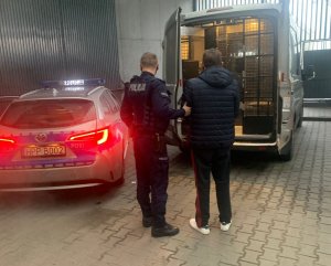 Umundurowany policjant trzymający zatrzymanego, który ma założone kajdanki na ręce trzymane z przodu. Widoczne na zdjęciu 2 samochody. Po lewej stronie oznakowany radiowóz.