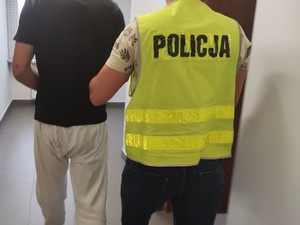 Policjant z założoną kamizelką z napisem POLICJA prowadzi zatrzymanego, który ma założone kajdanki na ręce.