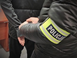 Zdjęcie przedstawiające policjanta zakładającego kajdanki na ręce trzymane z tyłu.