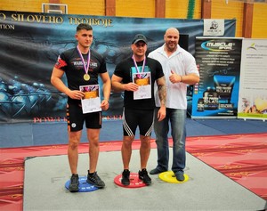Na zdjęciu 3 mężczyzn,stojących na podium podczas wręczenia nagród w zawodach uginania ramion ze sztangą.