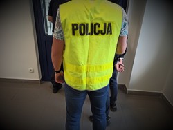 Na zdjęciu policjant kryminalny, który ma założoną kamizelkę z napisem POLICJA stoi z zatrzymanym mężczyzną w policyjnym areszcie.
