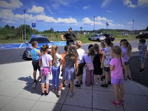 Na zdjęciu policjant prezentuje dzieciom tarczę policyjną i pałkę.