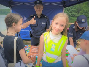 Na zdjęciu dziewczyna, która zdobyła odblaskową kamizelkę podczas jednego z konkursów. Za nią stoi uśmiechnięty policjant.