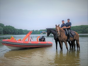 Na zdjęciu policjanci na koniach policyjnych stoją w wodzie przy motorówce strażackiej.