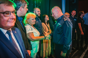 Na zdjęciu Komendant Powiatowy Policji w Będzinie składa gratulacje kobiecie na scenie, która stoi w rzędzie z innymi osobami.