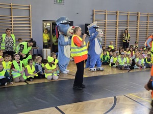 Na zdjęciu przemawiająca kobieta na sali gimnastycznej, za nią dwie maskotki policyjne.