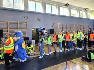 Zdjecie na sali gimnastycznej. Grupa osób dorosłych i policyjna maskotka w szeregu maszeruje w elementach odblaskowych.