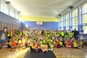 Zdjęcie grupowe na sali gimnastycznej uczniów, nauczycieli i policjantów podczas pokazu elementów odblaskowych.