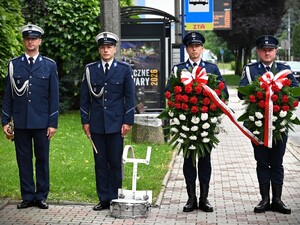 Na zdjęciu 4 policjantów stoi w szeregu. Dwóch z nich to policjanci z orkiestry Policji, a dwóch trzyma  biało-czerwone wiązanki kwiatów w rękach.