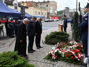 Dwóch mężczyzn w garniturach oraz ksiądz składają kwiaty pod pomnikiem, przed budynkiem komendy.