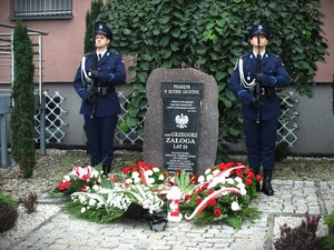 Na zdjęciu dwóch policjantów z warty honorowej stoją przy pomniku, przy którym złożono wiązanki kwiatów.