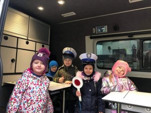 Pięcioro dzieci w policyjnym dużym radiowozie pozuje do zdjęcia, dwoje z nich ma założone policyjne czapki, a jeden trzyma tzw. lizak do zatrzymywania samochodów.