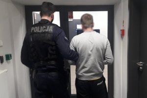 Umundurowany policjant prowadzi zatrzymanego mężczyznę korytarzem komendy.