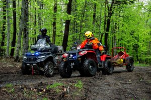 Policjanci jadą policyjnym quadem przez leśny teren razem ze strażakami, którzy również prowadzą służbowego quada.