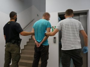 Nieumundurowani policjanci prowadzą zatrzymanego mężczyznę, który ma kajdanki założone na ręce trzymane z tyłu. Stoją przed wejściem do windy.