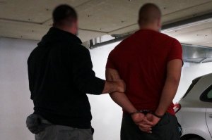 Nieumundurowany policjant trzyma zatrzymanego mężczyznę z założonymi na rękach z tyłu kajdankami. Zdjęcie wykonane przy nieoznakowanym radiowozie.