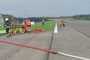 Na płycie lotniska stoi awionetka, obok pojazdy służb ratowniczych. Zdjęcie wykonane w dzień