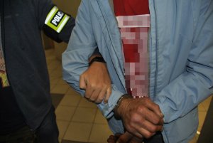 Zbliżenie na ręce zatrzymanego mężczyzny, na których są kajdanki. Pod rękę zatrzymanego trzyma nieumundurowany policjant, od którego widać fragment ręki z opaską z napisem POLICJA