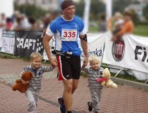 Podkomisarz Dariusz Stypułkowski podczas zawodów, biegnie trzymając za ręce swoich synów. Chłopcy trzymają pluszowe misie