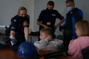 Policjantka prezentuje sprzęt policyjny dzieciom, które siedzą za stołem. Nad rodzeństwem stoi ojciec i policjant