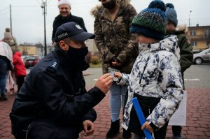 Policjant w granatowym mundurze klęczy przy małym chłopcu i wręcza mu odblaskową zawieszkę