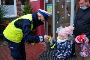 Przed wejściem do przedszkola policjant ruchu drogowego w odblaskowej kamizelce pochyla się nad dzieckiem wręczając mu odblask. Obok dziecka stoi mężczyzna