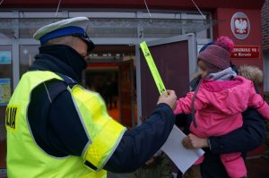 Przed wejściem do przedszkola policjant ruchu drogowego w odblaskowej kamizelce trzyma w uniesionej ręce odblaskową opaskę. Przed policjantem stoi dorosła osoba trzymająca na ręku małe dziecko