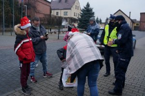 Na wybrukowanym placu stoi grupa osób, wśród których są policjanci i kobieta wręczająca dzieciom odblaski i słodycze