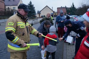 Umundurowany strażak wręcza dzieciom odblaskowe opaski. Na drugim planie stoi grupa dzieci