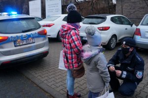 Na parkingu policjant w granatowym mundurze kuca przed dwójką dzieci wręczając im odblaski. Po lewej stronie stoi radiowóz, w tle zaparkowane samochody