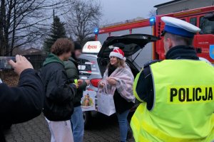 Na pierwszym planie tyłem stoi policjant ruchu drogowego w odblaskowej kamizelce z napisem POLICJA na plecach. Na drugim planie dziennikarka wręcza dwóm młodym chłopcom odblaski i słodycze. W tle widać wóz strażacki i samochód radia Express