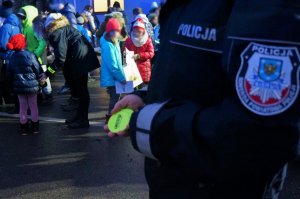 Na pierwszym planie fragment ręki umundurowanego policjanta, który trzyma odblaskowe zawieszki. W tle widać grupę dzieci