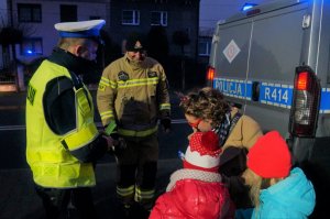 Przy radiowozie policyjnym stoi policjant ruchu drogowego i strażak. Przy nich kobieta pochyla się przy dwojce dzieci, częstując je słodyczami.