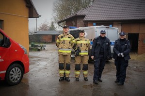 Na zdjęciu dwoje strażaków i dwoje policjantów pozują do obiektywu stojąc w szeregu. Z tyłu za nimi stoi policyjny radiowóz