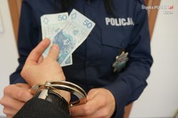 W dłoni trzymane banknoty, które podawane są policjantowi, który zakłada kajdanki