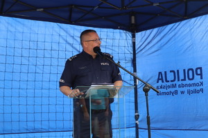 Na zdjęciu oficer policji stojący przed mikrofonem, pod niebieskim namiotem, będący w trakcie przemowy